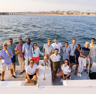 A group of colleagues smile for a group photo during team building activities in Orange County, standing together on the back of a whale watching boat overlooking Balboa Pier.