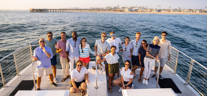 A group of colleagues smile for a group photo during team building activities in Orange County, standing together on the back of a whale watching boat overlooking Balboa Pier.