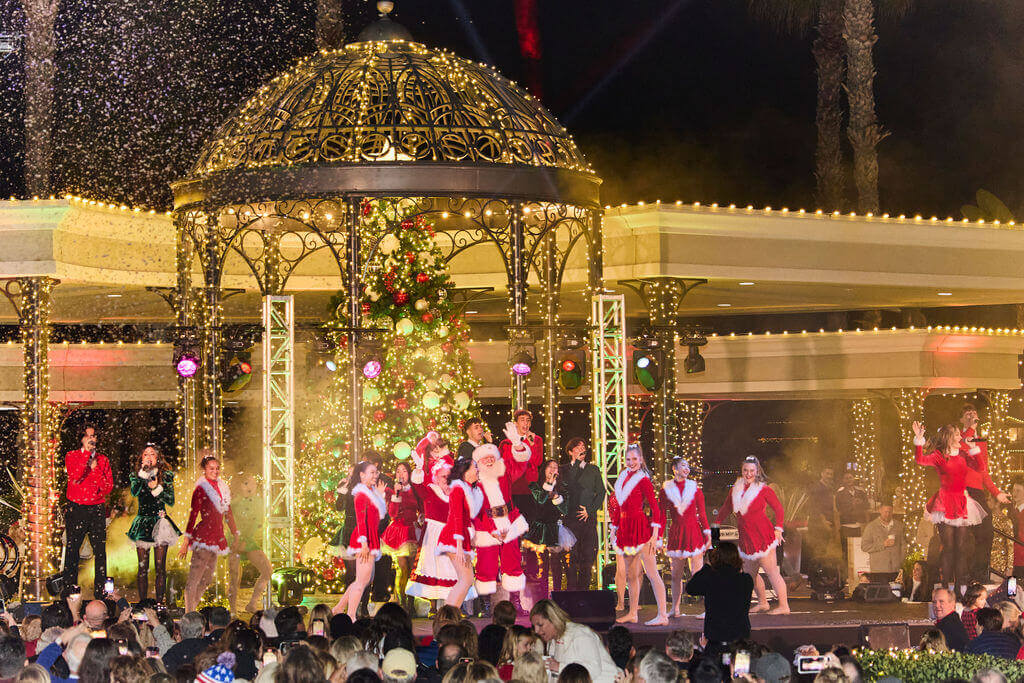 Christmas-themed dancers and vocalists wearing red and white, including Santa and Mrs. Claus, performing at Balboa Bay Resort's tree lighting celebration.