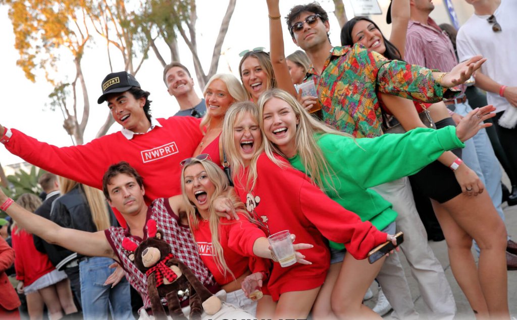 A group of young men and women wearing red and green Christmas-themed clothing, smiling and holding their arms out wide.